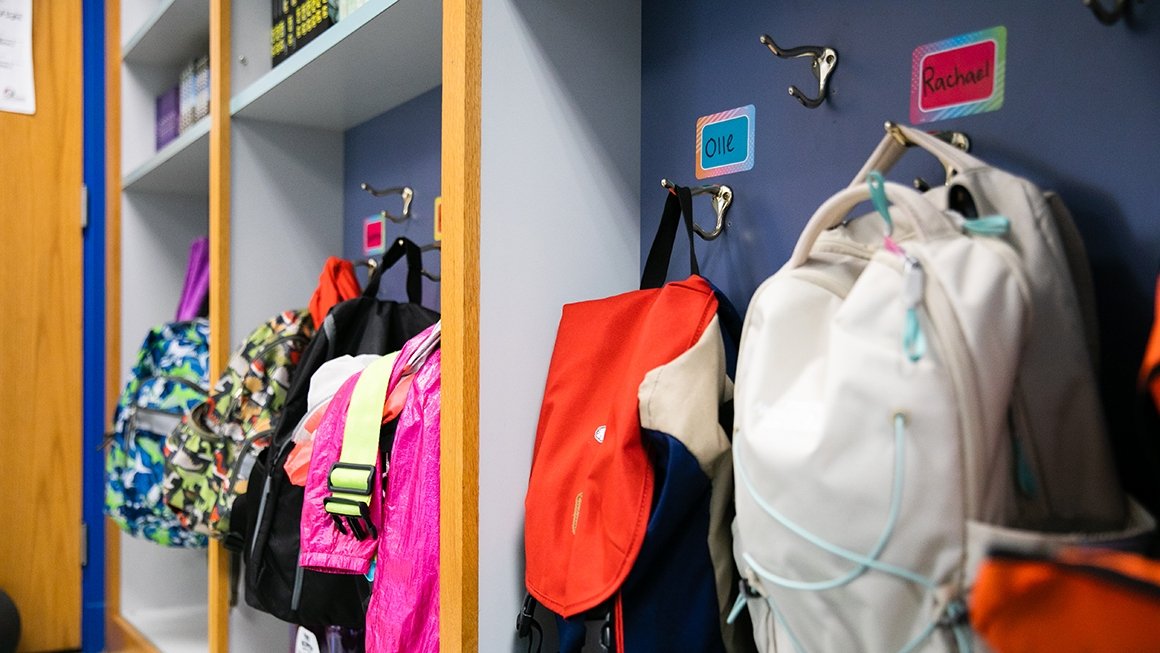 Backpacks hang on hooks with children's names printed on tags above each hook