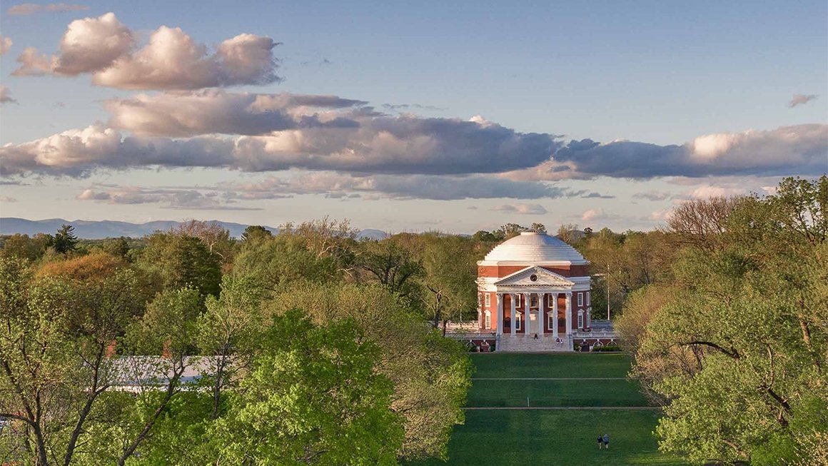 Aerial image of the UVA Rotunda and lawn in the spring surrounded by green trees and a cloudy blue sky