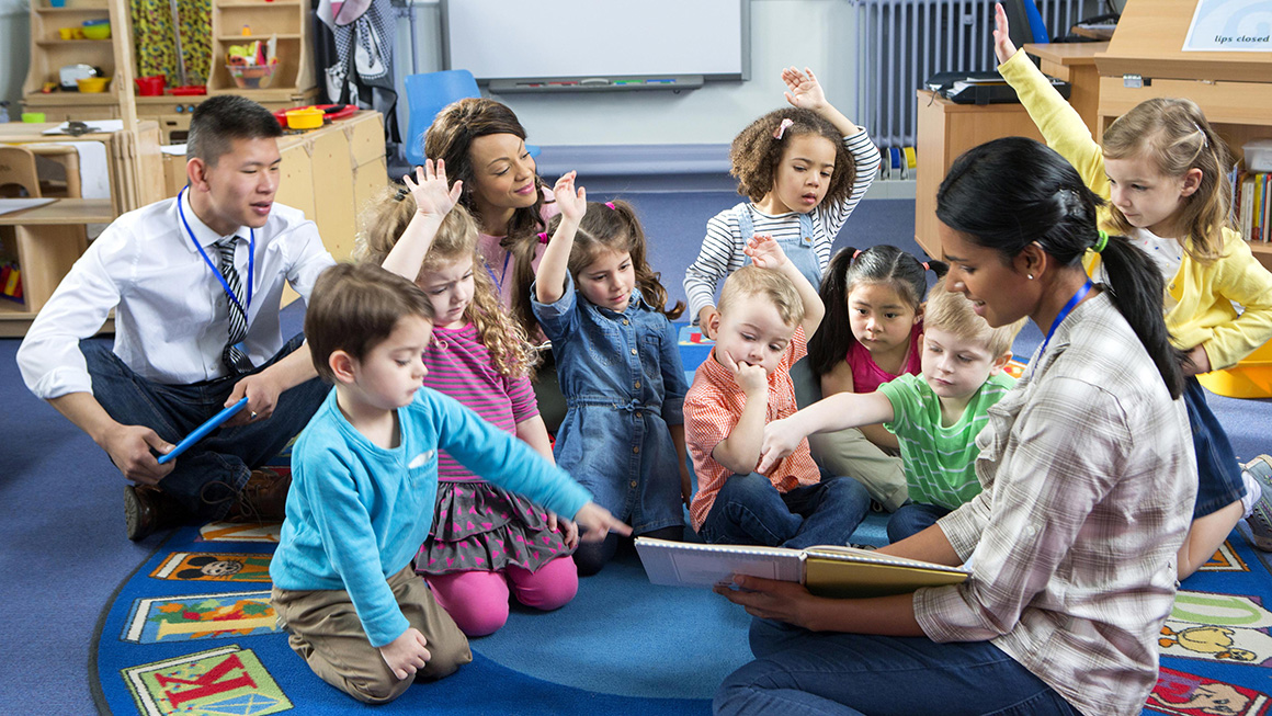 Preschool teacher reads to her class, all sitting on the carpet.