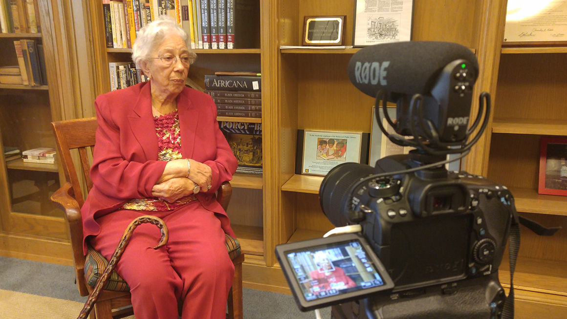 Ms. Spurlock sits in a chair wearing a read suit looking at a video camera