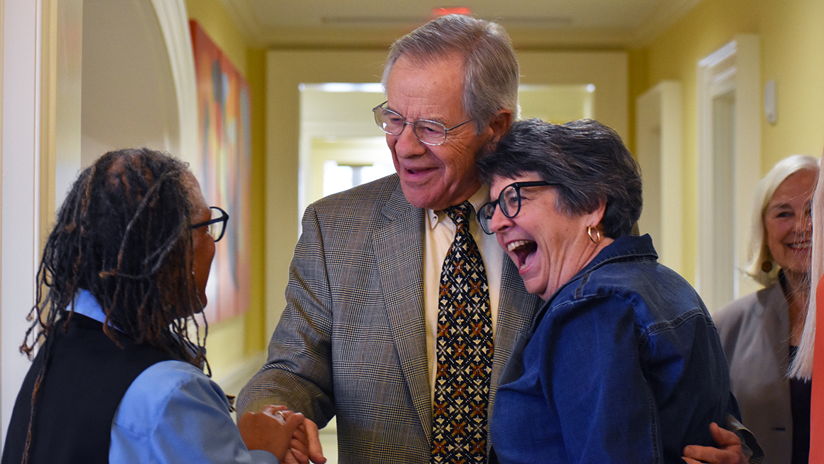 An older man wearing a suit shaking hands with one person while side hugging another person