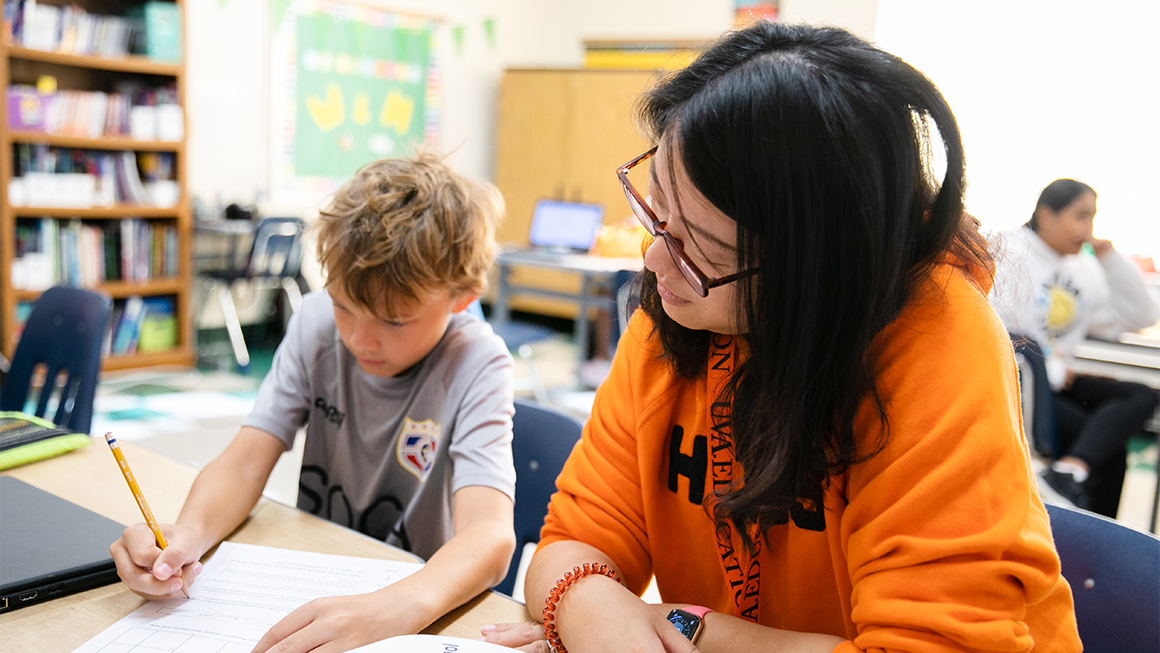 news_study-knowledge-rich-curriculum-boosts-reading-scores_1160x653_2023-04-12.jpg A teacher wearing an orange sweatshirt sits next to a young boy at a desk and looks on as he works on a worksheet.