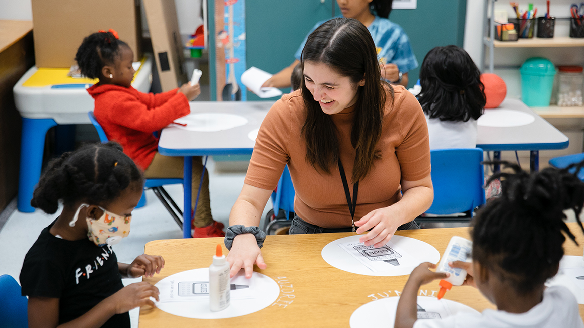 Teacher, smiling, sits at table with two students, while they work on a craft with glue.