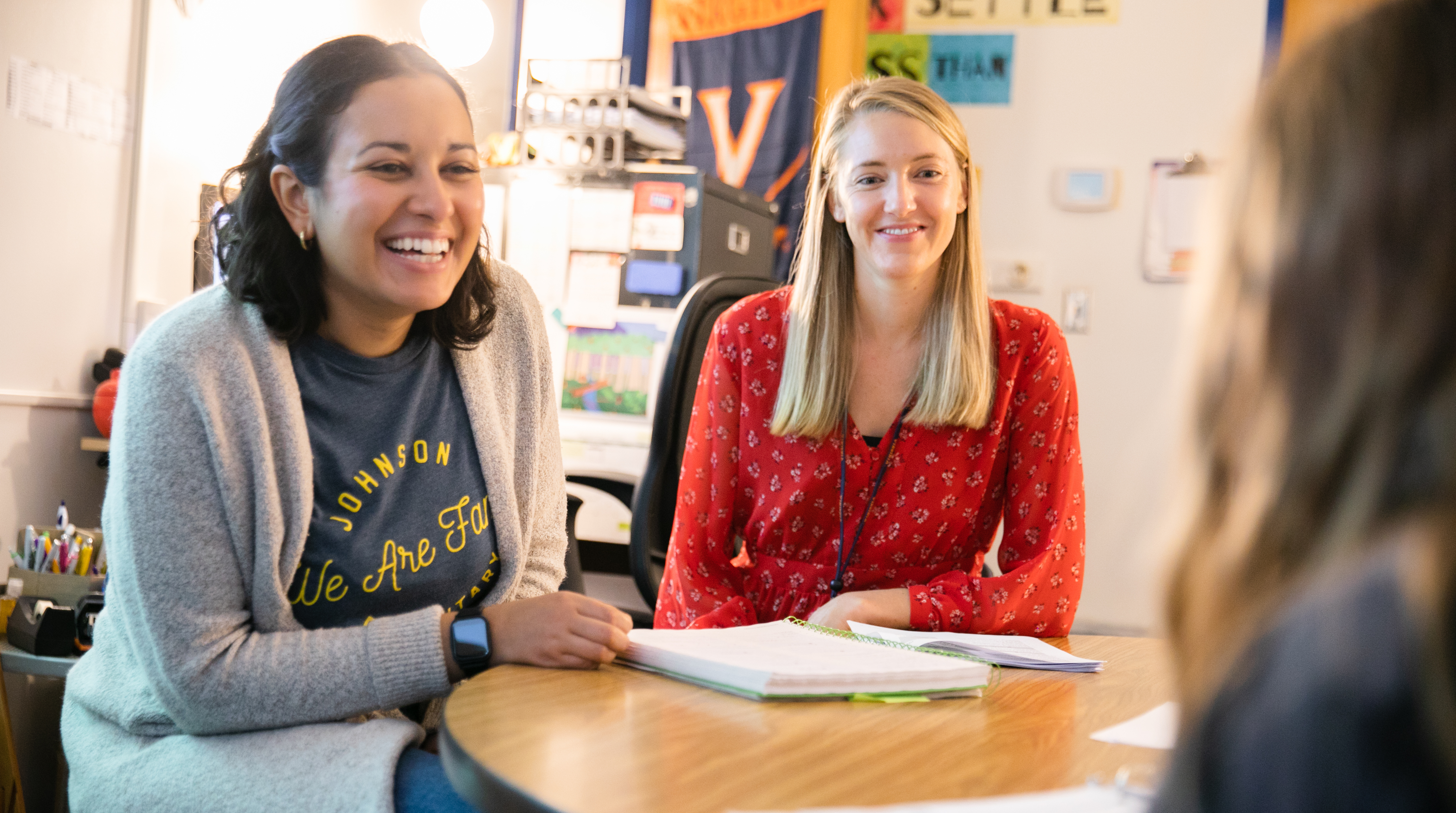 Three teachers gathered around a table. Two teachers are facing the third. They are smiling and have notebooks on the table.