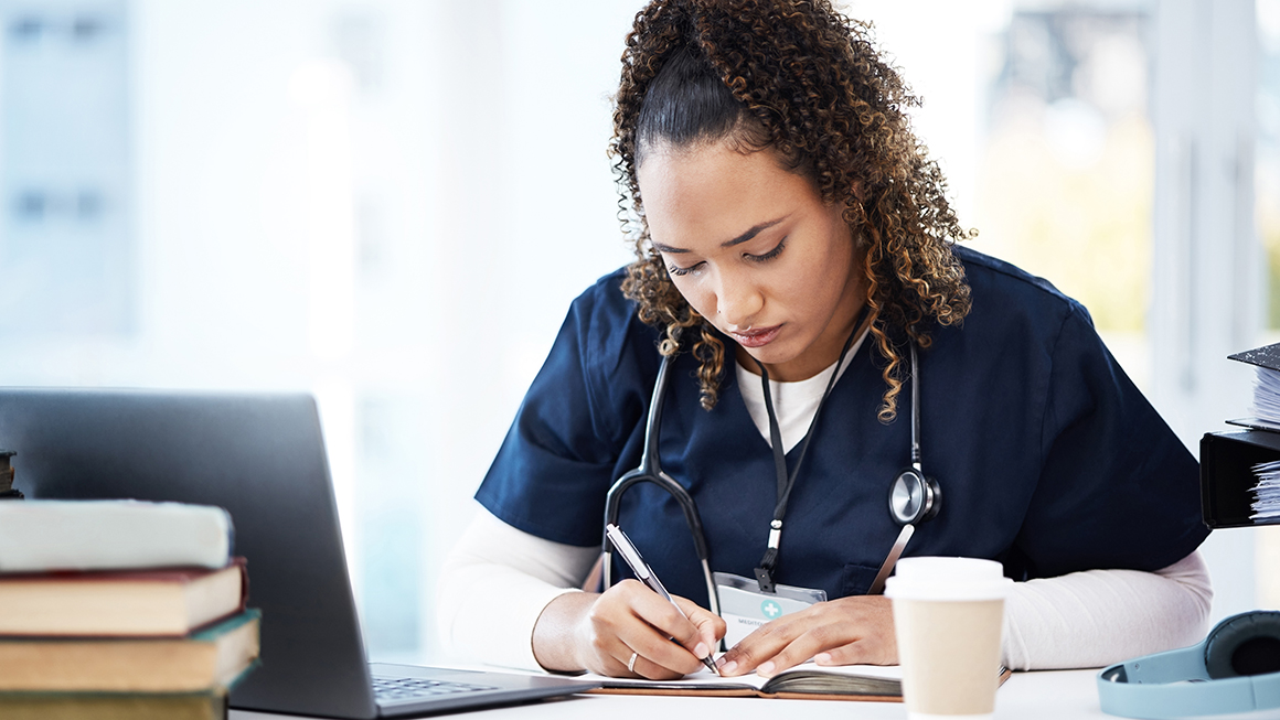 Woman wearing scrubs and a stethoscope sits in front of a laptop and writes in a notebook.