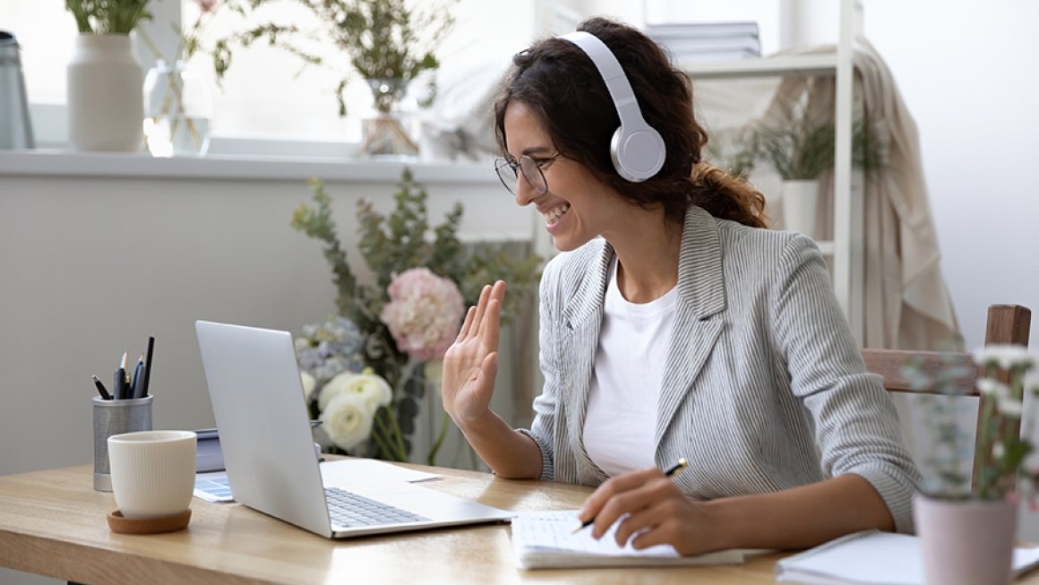 Woman sitting at desk wearing headphones, smiles and waves at open laptop screen.