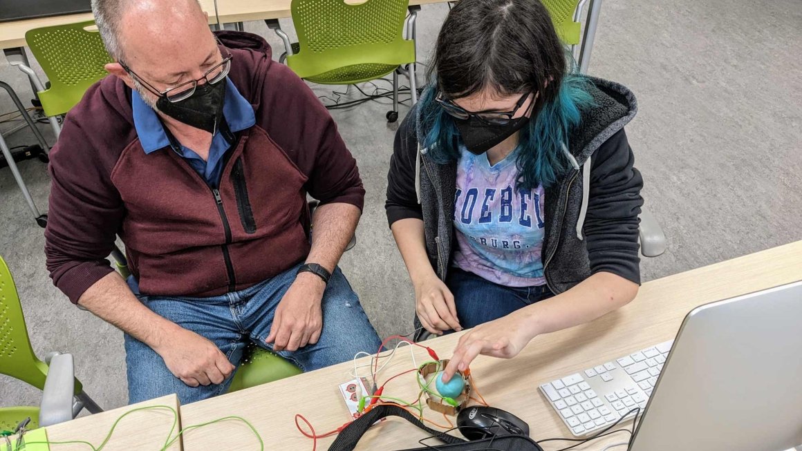 An older man and a young woman wearing masks sit at a table working on a computer science project together