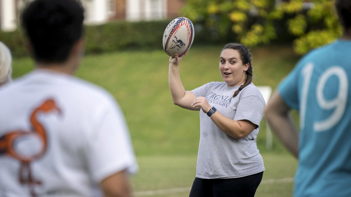 A woman standing on an athletic field wearing a gray T-shirt holds up a rugby ball as she speaks to other people on the field