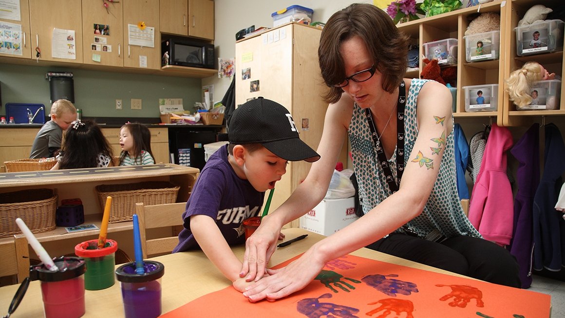 Teacher presses young boy's hand onto paper to make a handprint.
