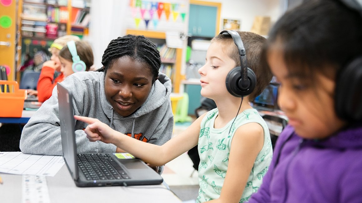 Teacher works with student while student wears headphones and touches screen of a laptop