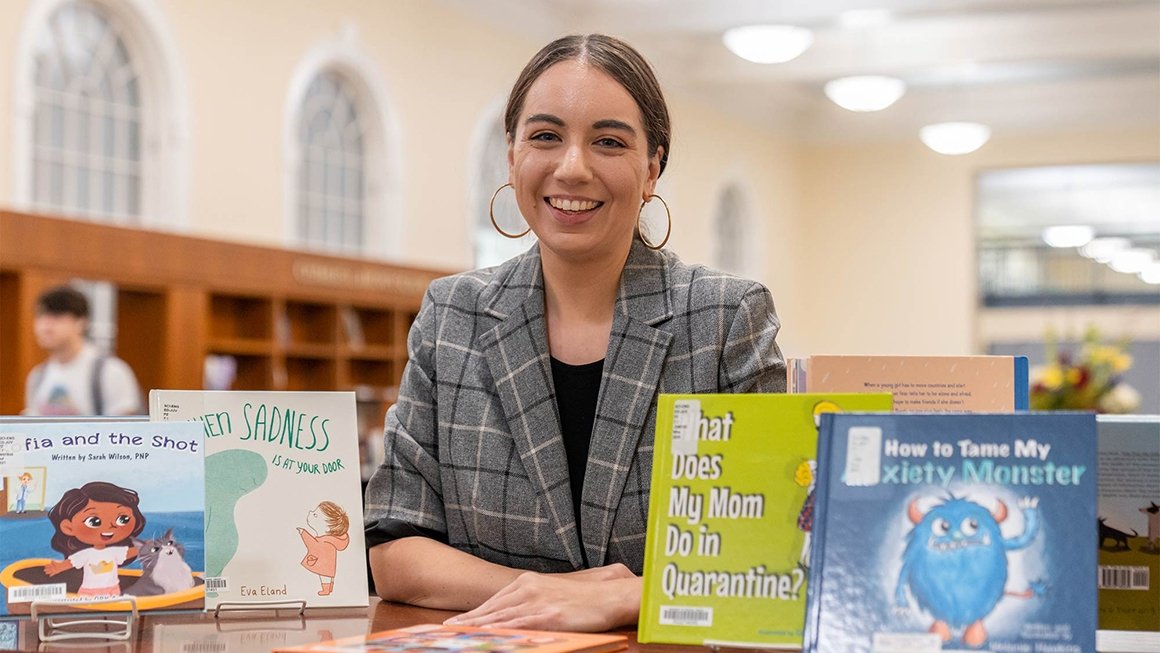 Ashley Hosbach stands behind bookshelf with children's books displayed on top.