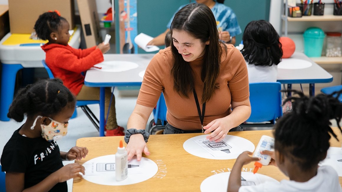 Teacher sits at table with two students doing a craft with glue