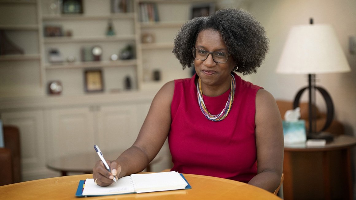 Stephanie Rowley wearing a red top sits at a round table writing in a notebook