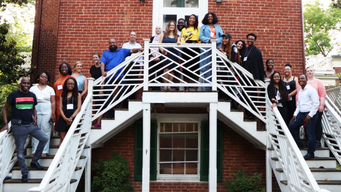 A group of undergraduate and graduate students stand on a white staircase outside of a brick building