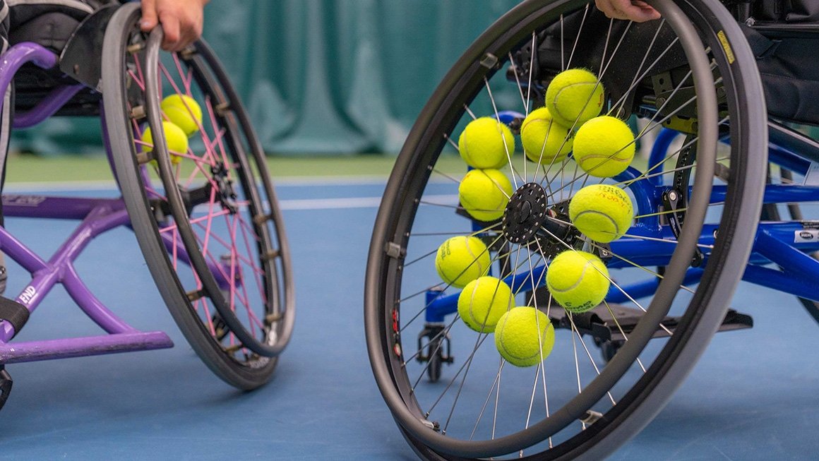 A close-up of two wheelchairs on a tennis court. Several tennis balls are stored in the spokes of the wheels.