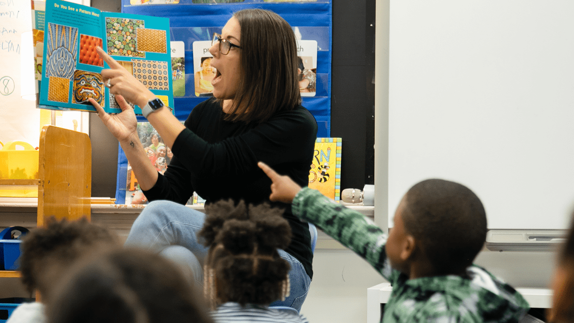 A teacher at the front of a classroom reading a book to students