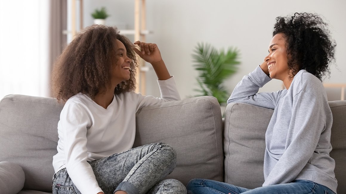 Adolescent and adult sit on a couch facing each other and smiling