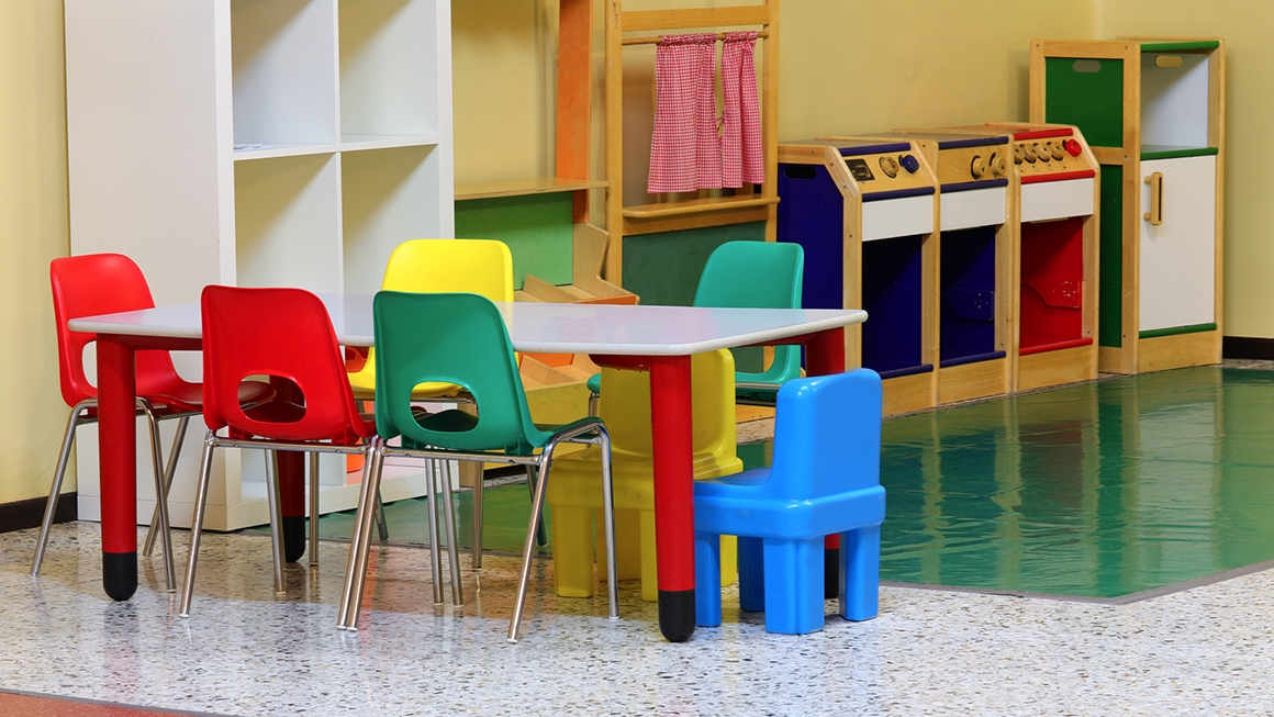Empty chairs of multiple colors surround empty table in preschool classroom.