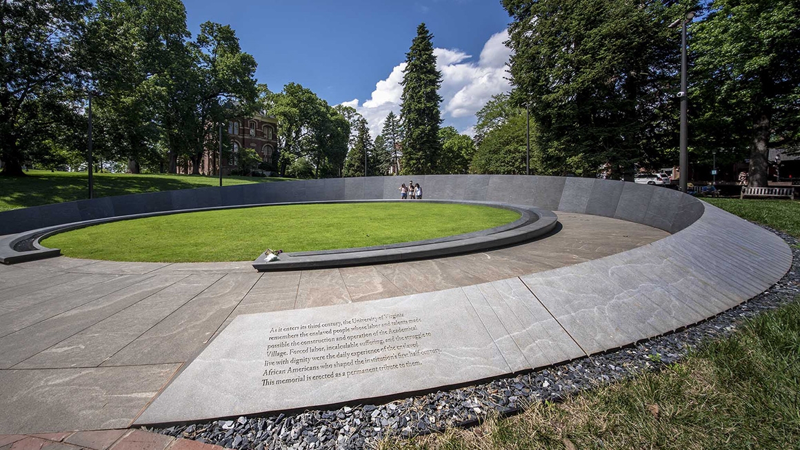 Three students stand in the center of a large circular concrete memorial surrounded by grass and trees. An inscription in the forefront reads: "As it enters its third century, the University of Virginia remembers the enslaved people whose labor and talents made possible the construction and operation of the Academical Village. Forced labor, incalculable suffering, and the struggle to live with dignity were the daily experience of the enslaved African Americans who shaped the institution's first half century