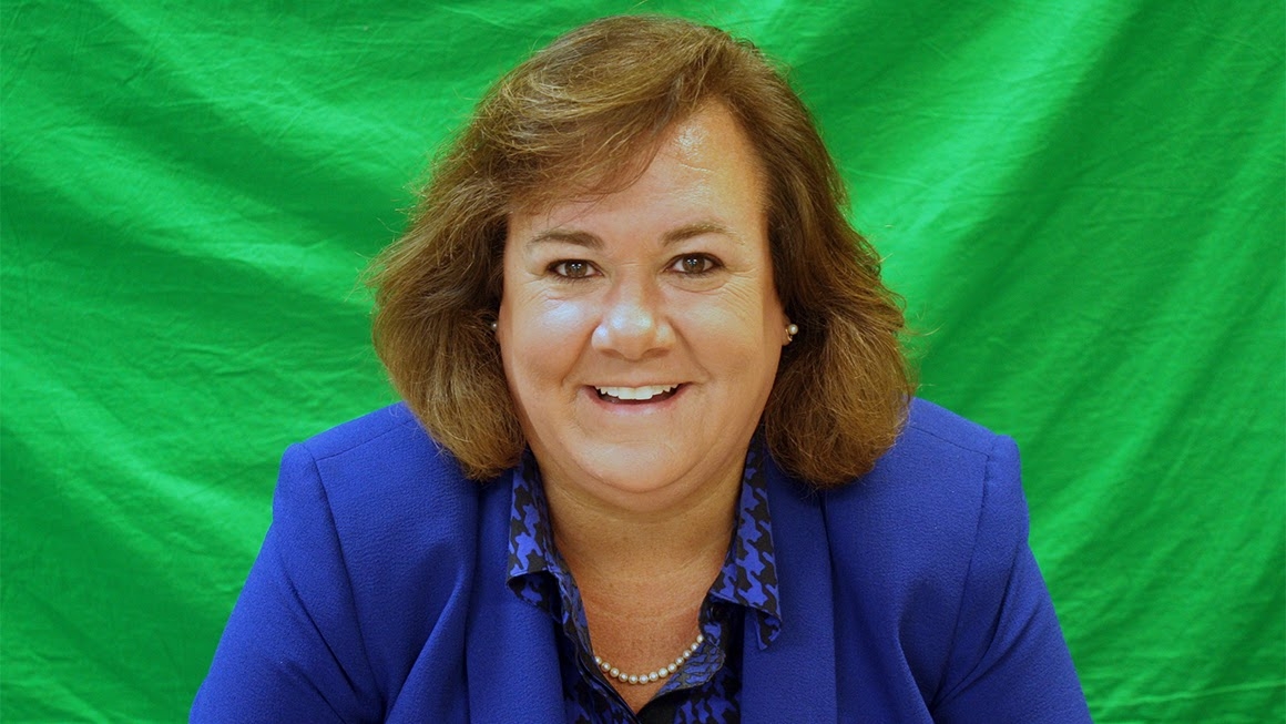 Mahri Aste wearing a blue jacket in front of a bright green background smiles at the camera