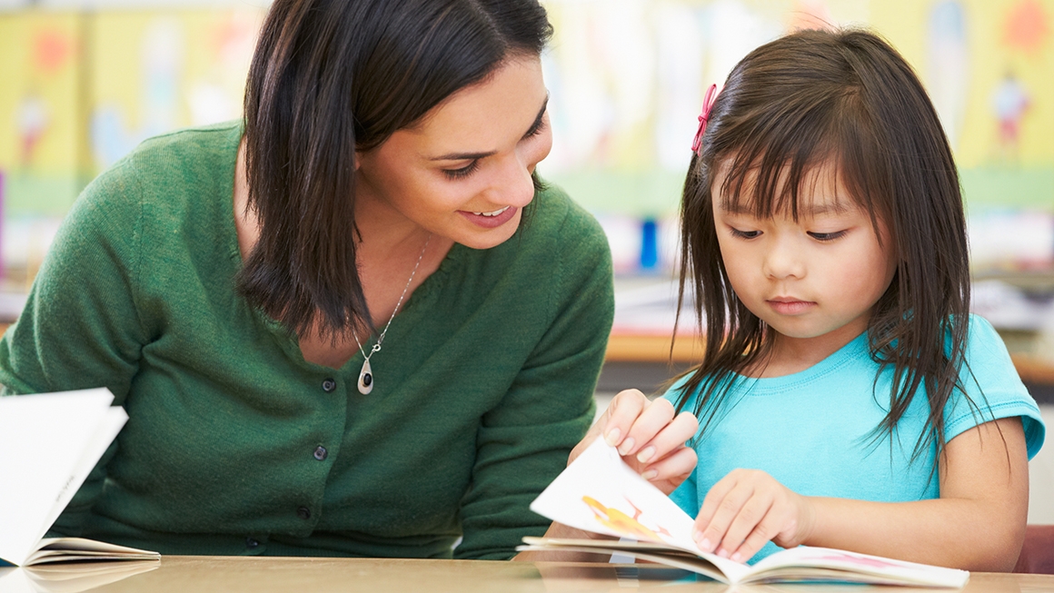 Woman sits at table next to young girl and they both look at open book.