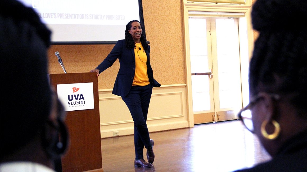 Bettina Love, wearing a yellow top and a black blazer, stands next to a podium and speaks to a crowd