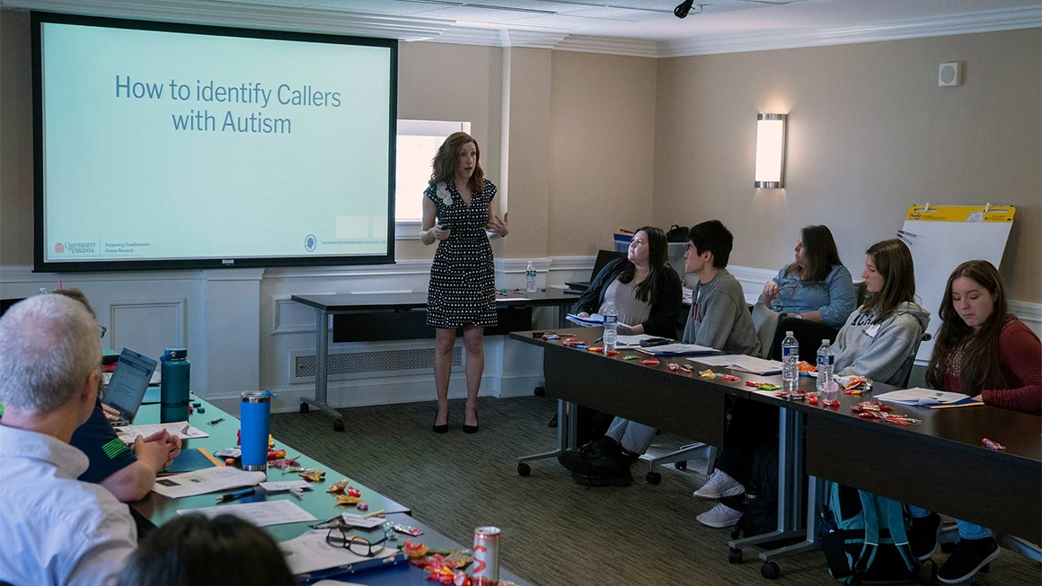 A woman stands in front of a screen that reads, "How to identify callers with Autism" while speaking to about 15 people seated at tables
