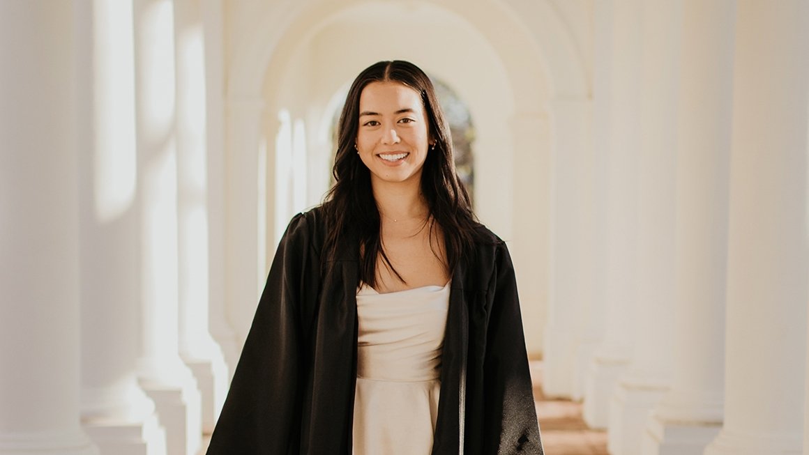 Sara stands in a colonnade wearing a graduation gown, smiles at the camera