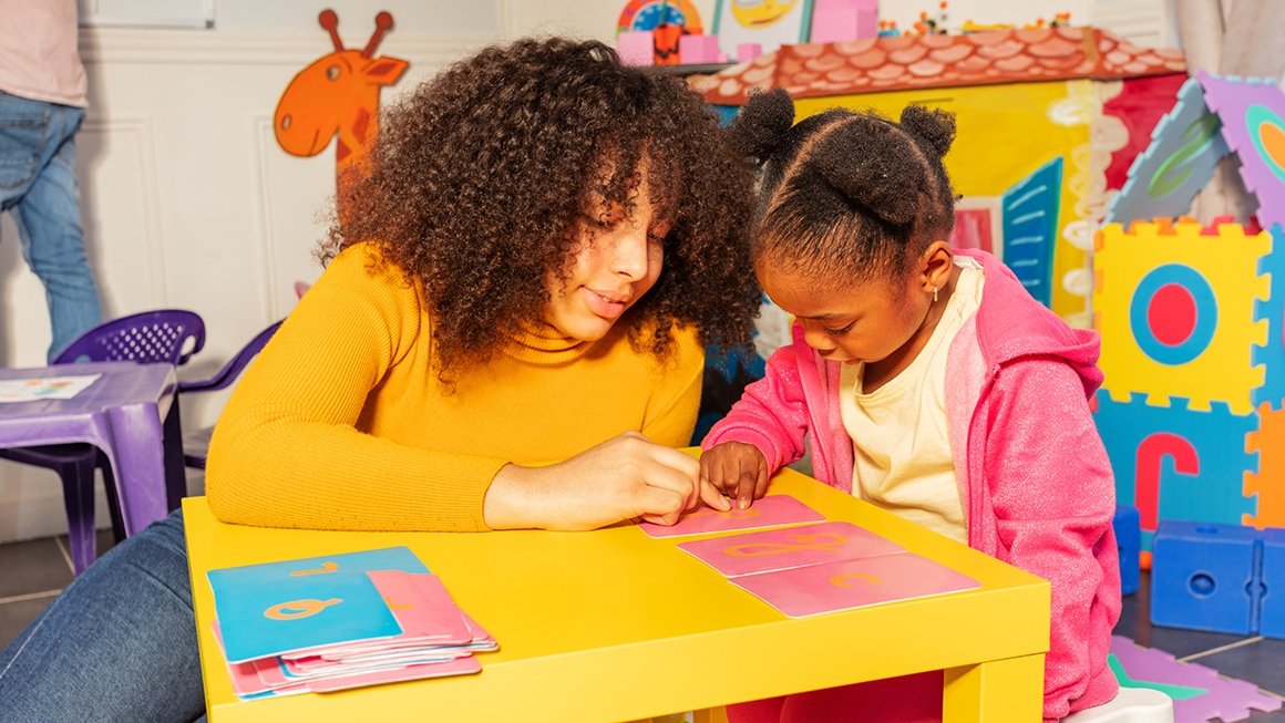 Teacher sits at small table beside a preschool student