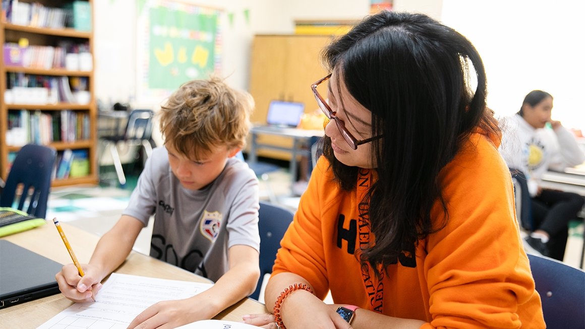 A teacher wearing an orange sweatshirt sits next to a young boy at a desk and looks on as he works on a worksheet.