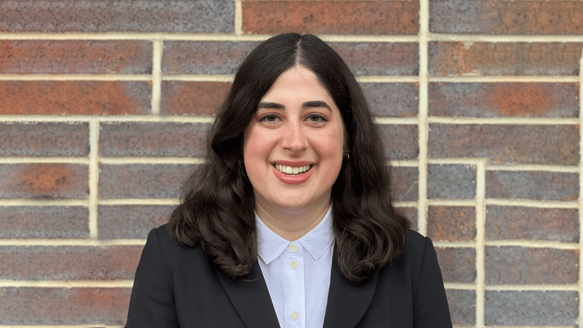 Lauren Coury, smiling and wearing a white button-down shirt and a black jacket, stands in front of a brick wall