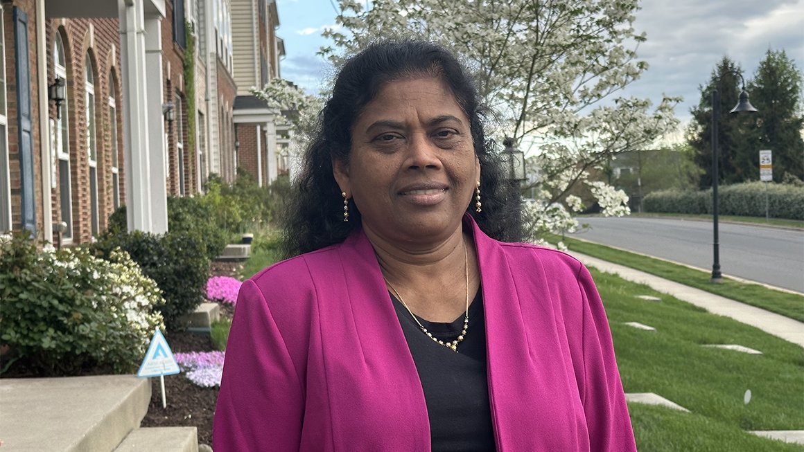 Lydia Justus, wearing a black top and bright pink jacket, stands outside among green grass and flowering trees.