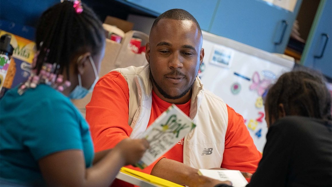 Quan Dennis, wearing an orange shirt with a tan vest, reads books with two young students in an elementary school
