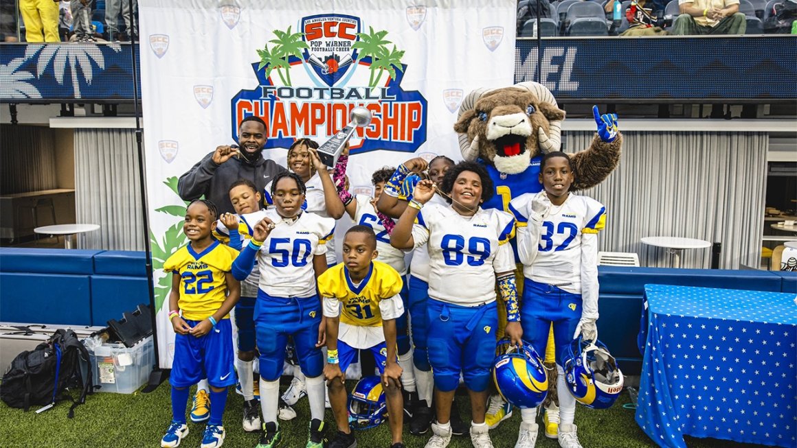 A group of middle school-age boys, wearing football uniforms in the bright blue and yellow of the Los Angeles Rams, poses on a football field in front of a Super Bowl banner with their team's director.