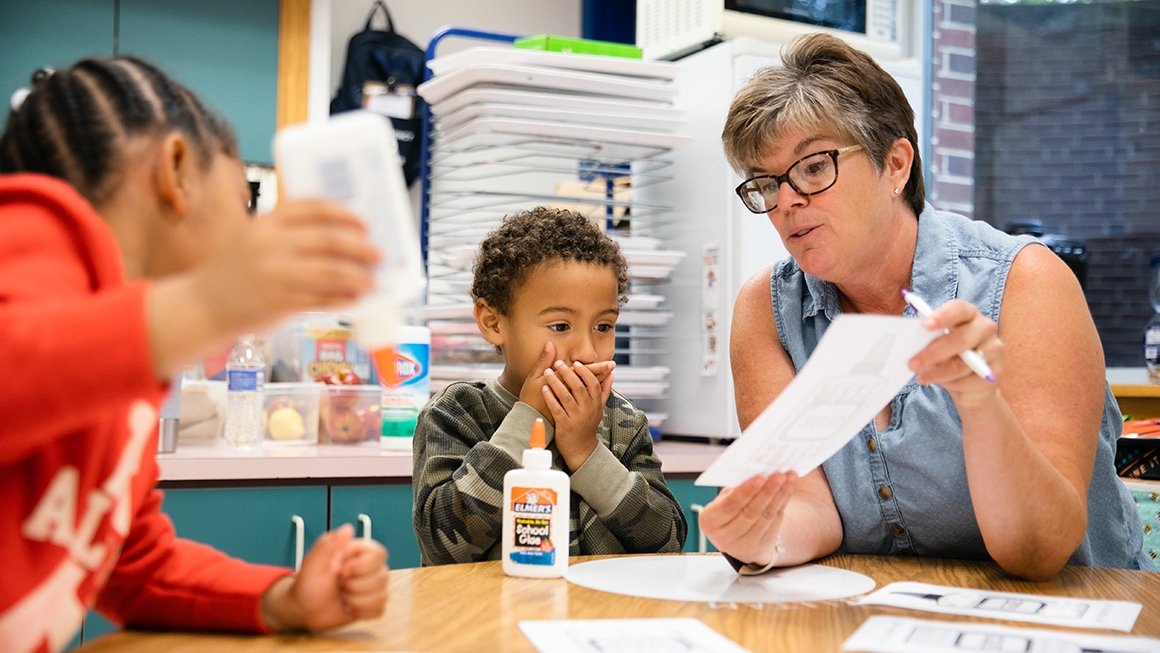 Teacher sits with two students showing them a paper. A boy student holds his hand over his mouth in surprise.