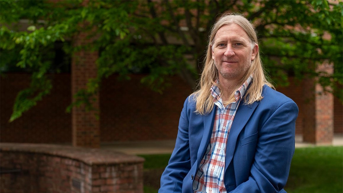 Chris Hulleman wearing a blue jacket sits outside against a backdrop of green trees and red bricks.