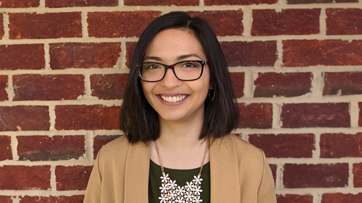 Alexis Prijoles, wearing a tan jacket, stands in front of a brick wall and smiles at the camera.