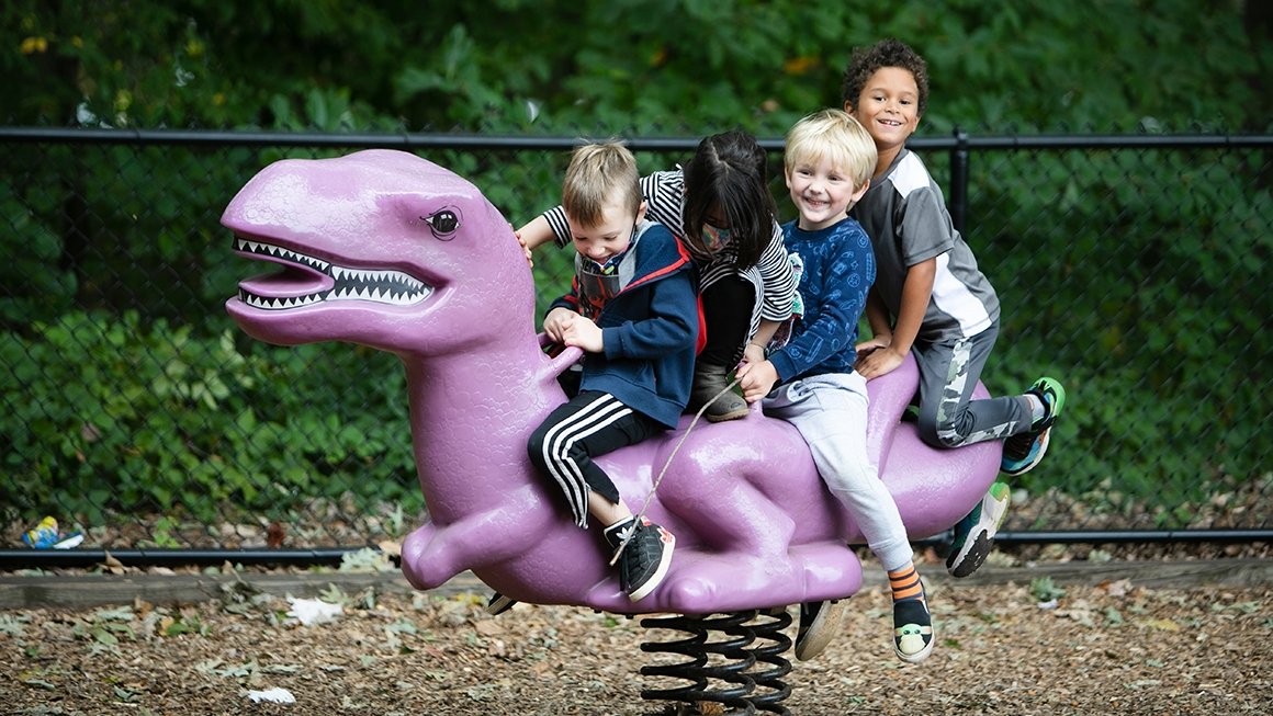 Four children ride a purple dinosaur on a playground. Two are looking down and two are looking at the camera and smiling.