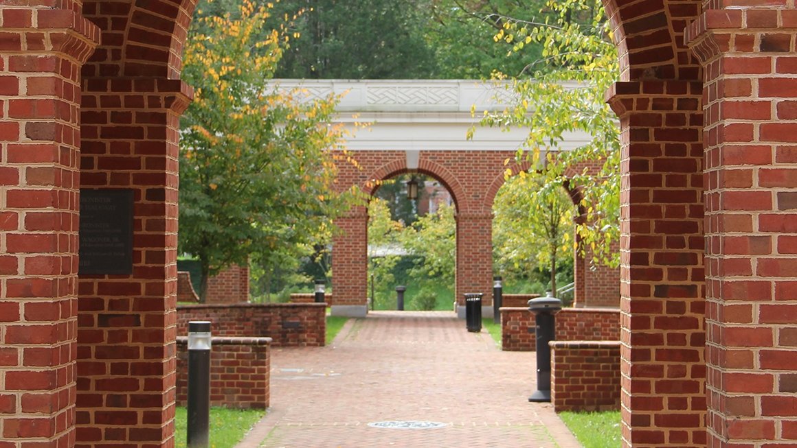 Trees frame a brick pathway. In the back is a walkway with arches.