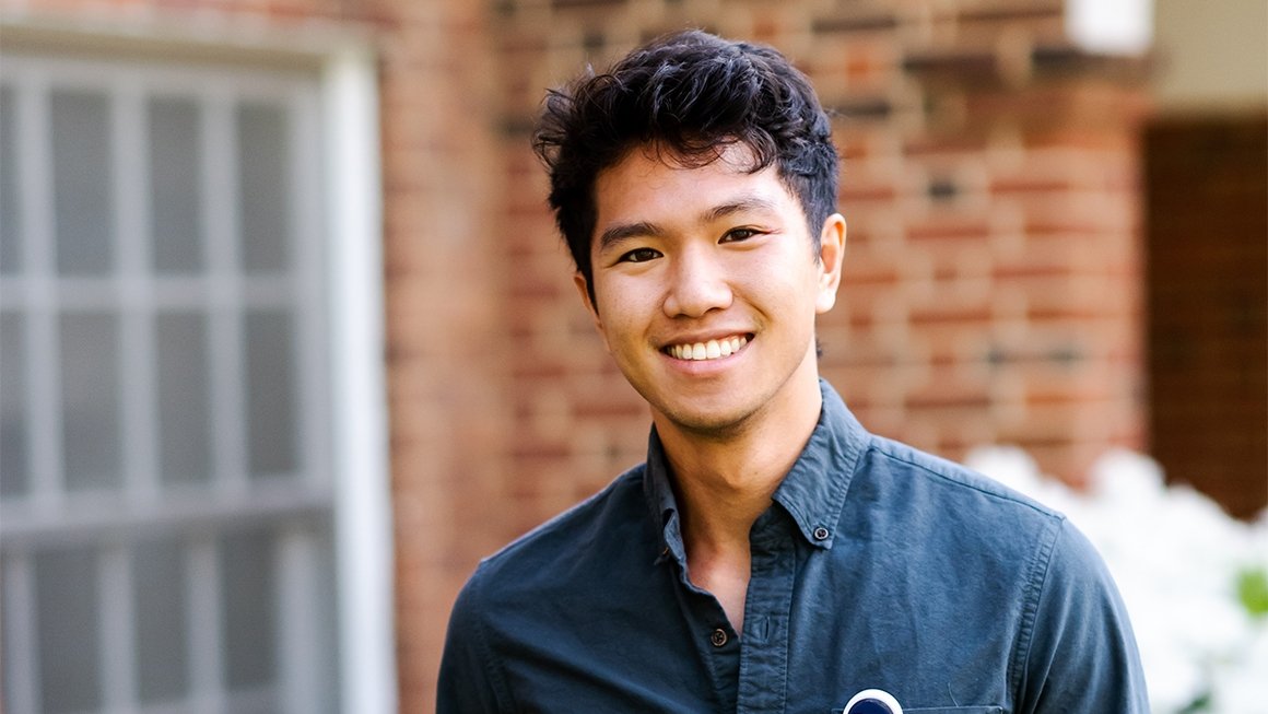 Jeison De Guzman wearing a dark button-down shirt stands outside in front of a brick building smiling at the camera