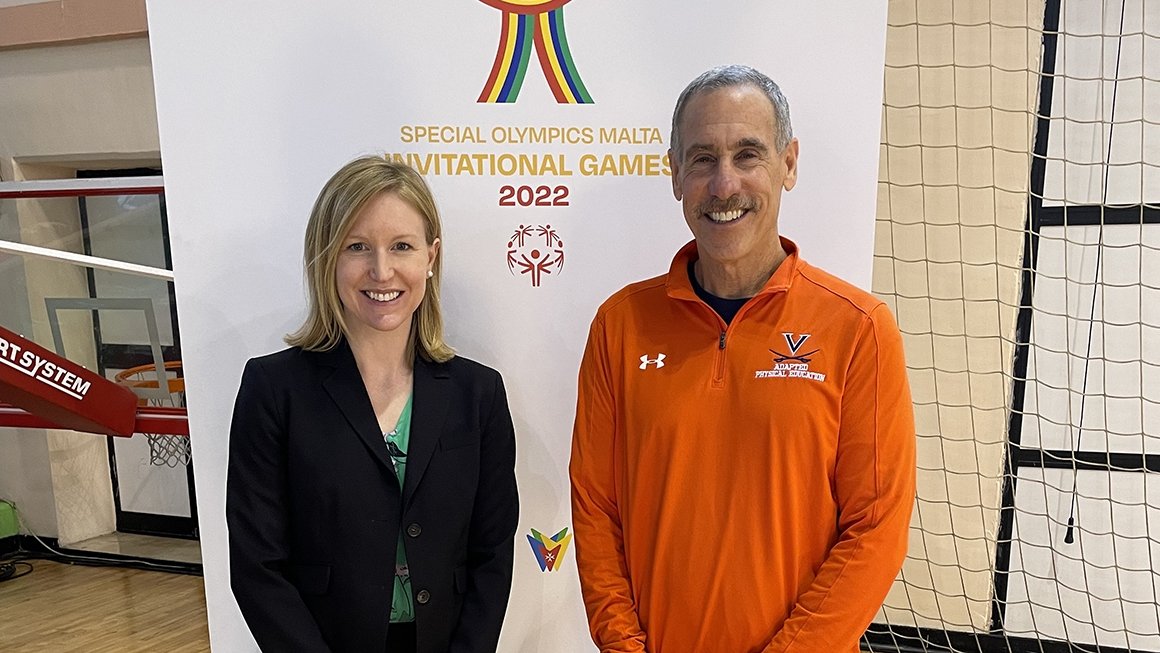 Martin Block, wearing an orange shirt with a UVA logo, stands beside a woman in a black suite in front a banner that reads Special Olympics Malta Invitational Games 2022