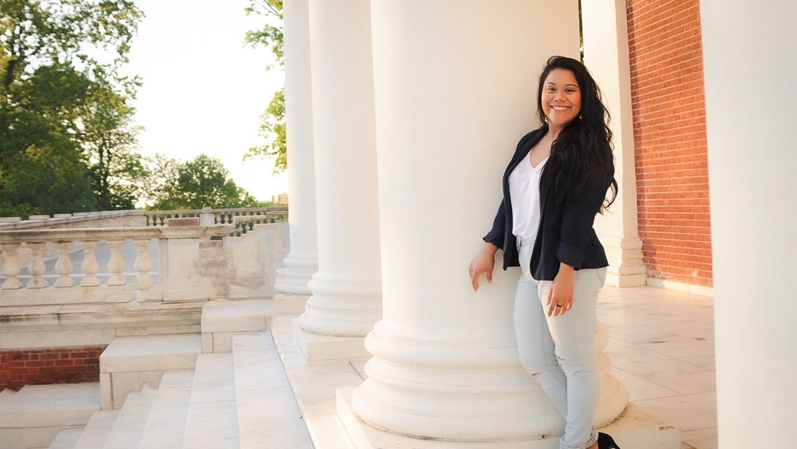 Bridget Drain, wearing jeans, a white top and a black blazer, leans against a pillar on the steps of the Rotunda at UVA