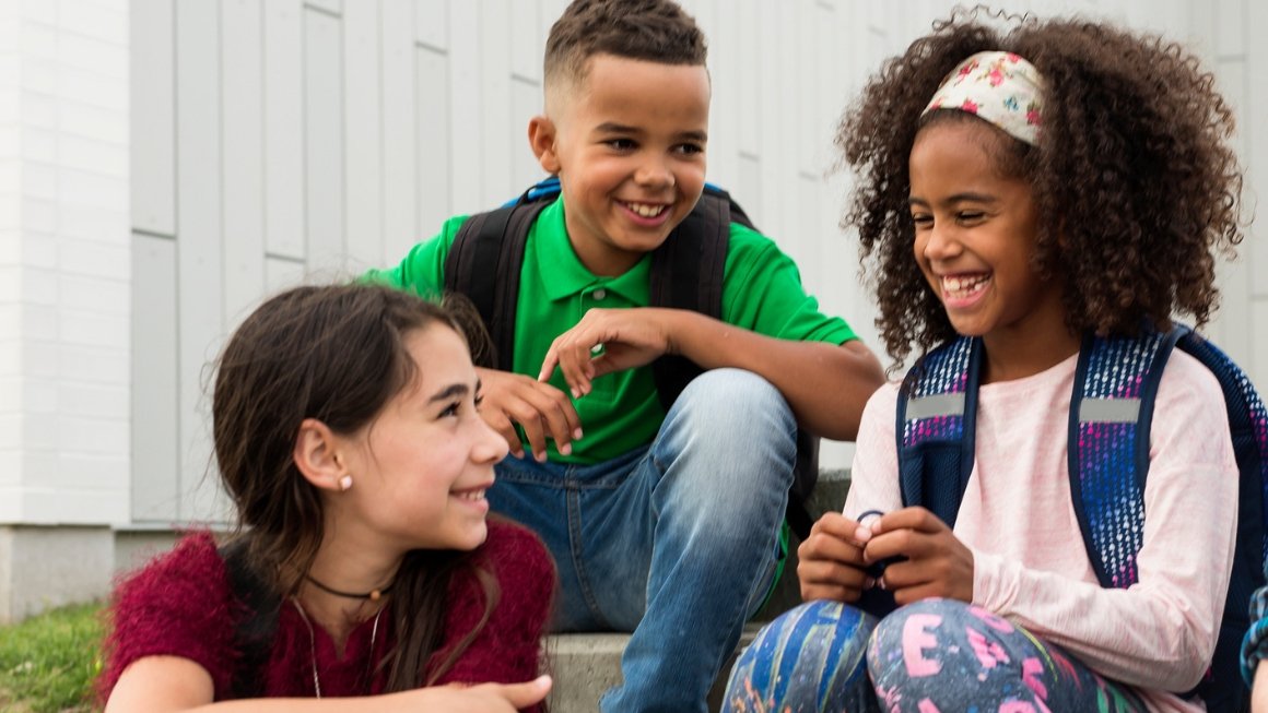 Three students sit on steps wearing backpacks, smiling.