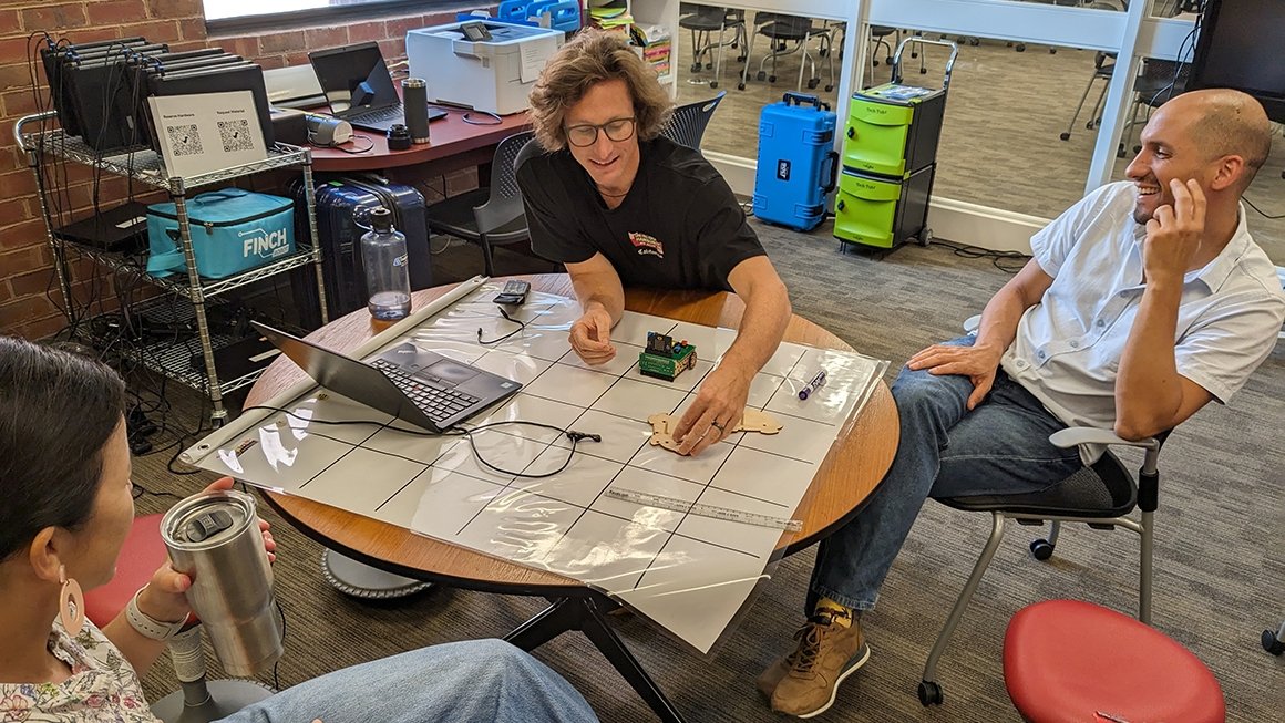 Three people sit around a table where small parts of a robot are sitting on a mat