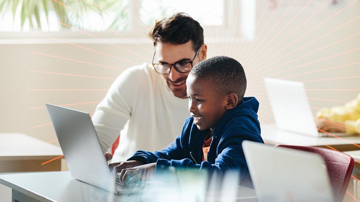 Teacher leans down next to young student working on a laptop at his desk.