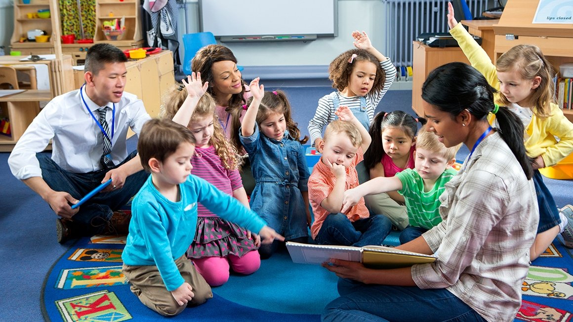 In a classroom, a teacher reads a book to students who are sitting together on the floor