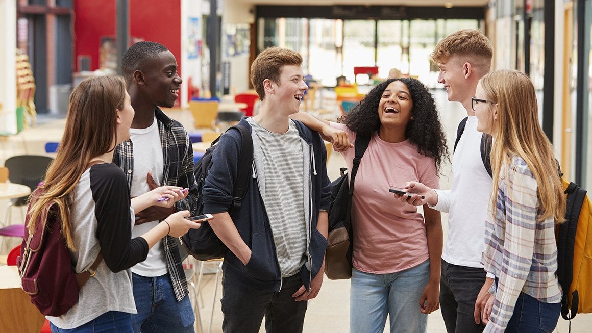 Youth stand in semi-circle laughing together. Some wear backpacks.