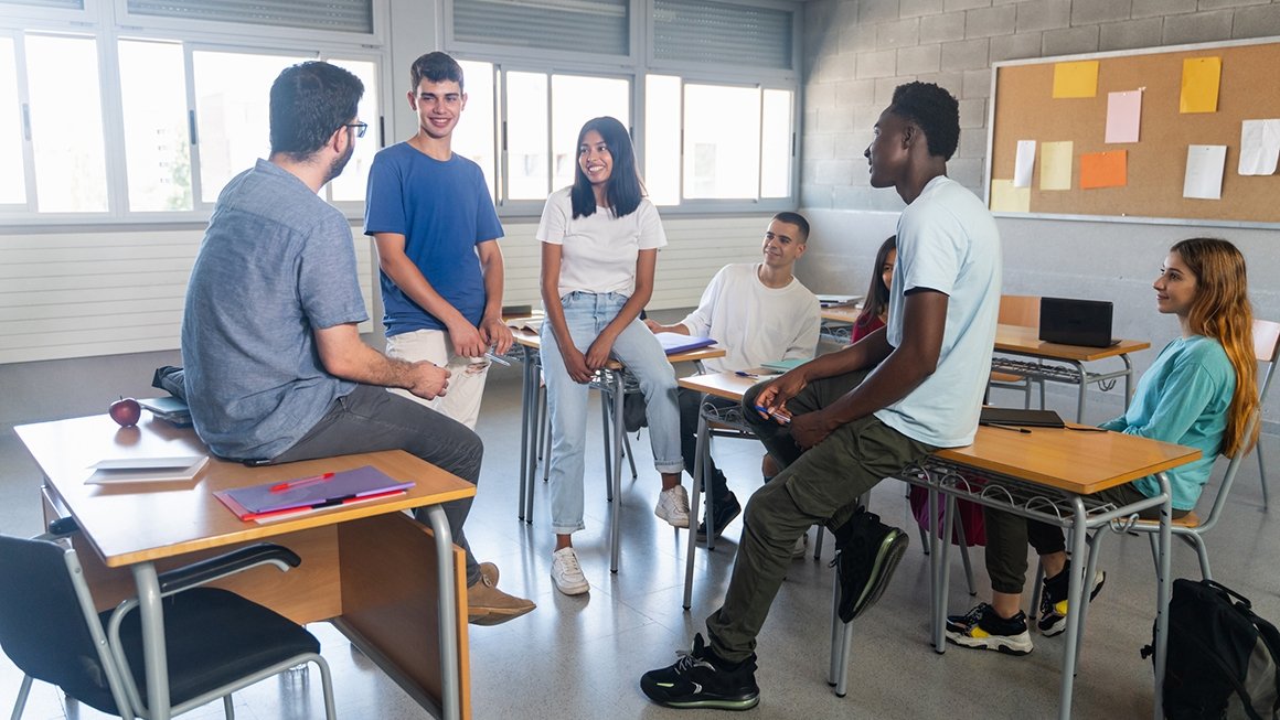 Students sit on desks in a circle and speak with teacher in a classroom