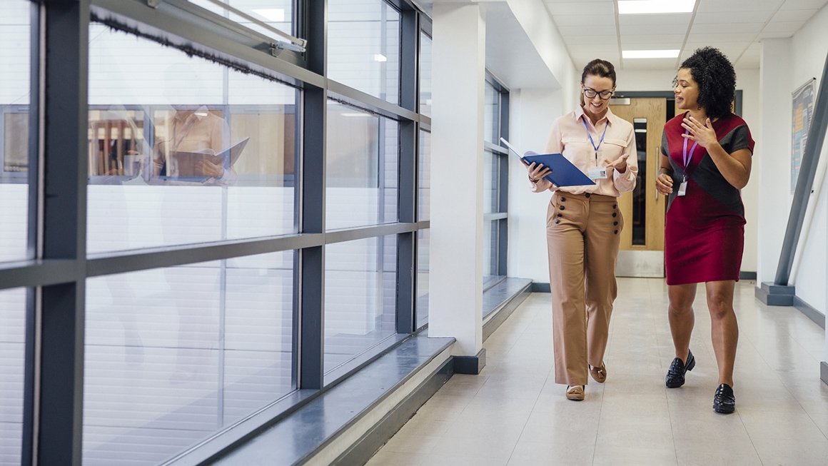 Two female teachers talk to each other walking down a hall in a school