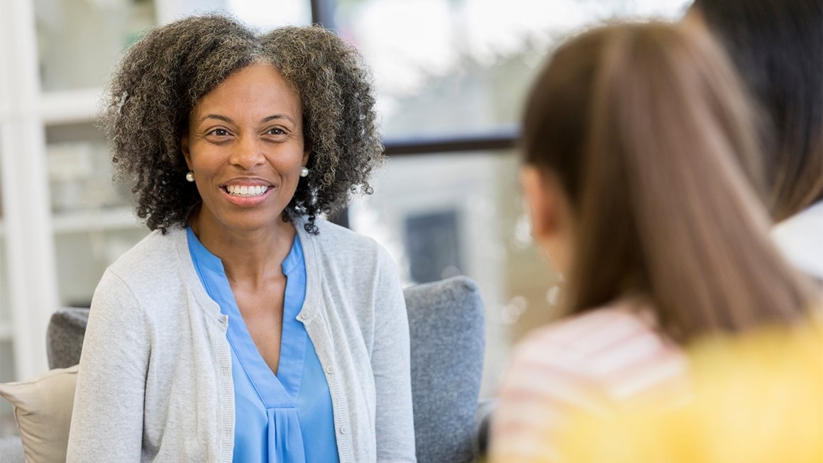 A woman wearing a blue shirt smiles while talking with a young woman 